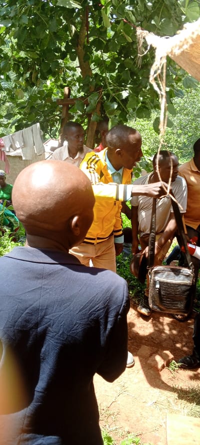 The image depicts an outdoor gathering of several men under the shade of a large, leafy tree in Kundu 1, Democratic Republic of the Congo. The scene is illuminated by bright daylight, casting strong shadows on the reddish-brown dirt ground, suggesting it is midday.

In the immediate foreground, a man with a bald head is seen from behind, wearing a dark blue suit jacket. His back is to the camera. Facing away from the foreground man, a central figure in a distinctive yellow and black striped athletic jacket is actively engaged, holding onto a thick piece of rope or twine that descends from the tree above. He appears to be speaking or demonstrating something to the group.

To the right of this man, another individual holds a dark, textured shoulder bag and a dark hat. Further in the background, other men are visible, observing the interaction. One man behind the central figure has a beard and is wearing a light-colored collared shirt. In the far left background, partially obscured by lush green foliage, a woman in a green outfit can be faintly seen, and clothes are hanging on a line near a simple building structure.

The setting is rural, characterized by dense green foliage, bare earth, and rustic surroundings. The people appear attentive and engaged in what seems to be a community meeting or a discussion. No discernible text is visible on any objects or clothing.
