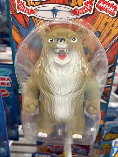 This is a close-up, eye-level photograph of a stretchy toy bear in its original packaging. The toy is light brown and white, with a furry texture. It has a wide mouth with visible teeth and red inner mouth details. The bear is enclosed in clear plastic packaging, which is attached to a colorful cardboard backing featuring a red arrow with Cyrillic lettering and an illustration of a person stretching. The packaging is displayed on a retail shelf, with other products visible in the background. The context of the image suggests it was taken in a store. The location is indicated to be Kuzmolovsky, Russia.