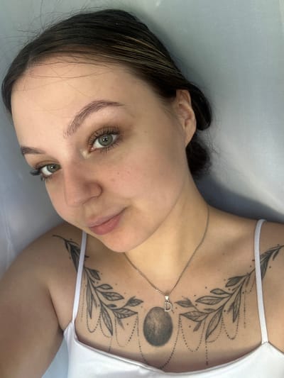 This is a close-up, high-angle, vertically oriented, eye-level shot of a young woman lying down. She is positioned in the lower right quadrant of the frame, with her face turned towards the camera. Her dark hair is partially obscuring her face, with some strands falling across her forehead and cheek. She has light skin and striking green eyes with dark eyelashes. Her lips are slightly parted and have a natural pink hue. She is wearing a white tank top, and a delicate silver necklace with a small, horseshoe-shaped pendant rests on her collarbone.
The woman has intricate black tattoos on her chest and upper chest area, featuring a branching vine with leaves and dotted lines. A circular design, possibly representing a moon or planet, is also visible as part of the tattoo. The overall tone of the image is soft and intimate, with a focus on the woman's features and the detailed tattoos. The lighting appears to be natural and diffused, suggesting an indoor setting. There is no visible text in the image.
