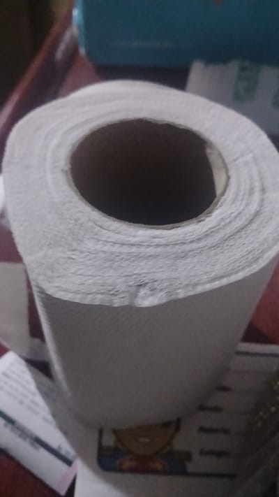 The image shows a roll of paper towels, likely in a household setting. The roll is mostly full, with a noticeable cardboard core visible in the center. The paper towels are white and appear slightly textured. Other objects are visible in the background, including what appears to be a blue container, a printed item, and possibly a tube. The precise location in Putucual, Venezuela, cannot be determined from this image.