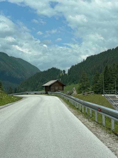 The image depicts a winding paved road in Flachauwinkl, Austria. The road curves to the left, bordered by a metal guardrail. To the right of the road, there is a small wooden structure with a sloped roof, resembling a shed or cabin. Beyond the cabin, a dense forest of evergreen trees covers the steep slopes of the mountains. In the background, layers of mountains recede into the distance under a cloudy sky. The scene is set in a natural, mountainous environment.