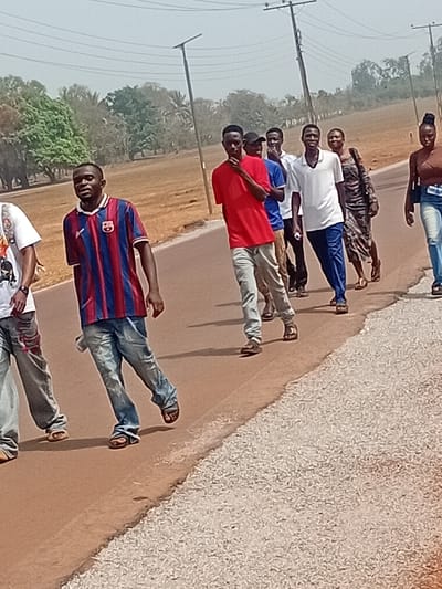 Eight men stand in a line along the edge of a road. Most are dressed in casual shirts and jeans. One wears a striped jersey, and another is shirtless. They appear to be posing or waiting together. Power lines and trees are visible in the background. The roadside is gravelly, contrasting with the paved road they are standing on. The scene is captured in a slightly skewed perspective.