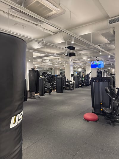 This is a photograph of the interior of a modern gym. The gym has a dark gray rubberized floor and numerous pieces of black gym equipment, including weight machines and a large punching bag on the left side of the frame. The ceiling is white and features exposed ductwork, lighting fixtures, and a large air conditioning unit. A flat-screen television is mounted high on the wall in the background, displaying a sports broadcast. The overall impression is one of a clean, well-lit, and functional fitness facility. The image does not provide any specific visual cues to determine its location within Kadıköy, Turkey.