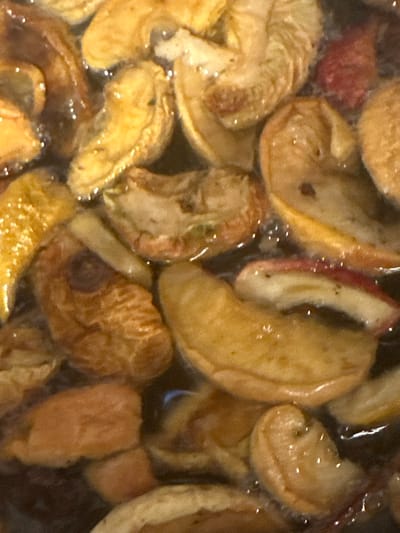 This close-up, high-angle shot captures a bowl of stewed apples in a dark, liquid medium, likely a pot or saucepan. The apples are sliced and appear to be in various stages of cooking, with some retaining their shape and others becoming soft and slightly broken down. Their colors range from pale yellow to golden brown, with hints of red skin visible on some pieces. The liquid is dark and murky, reflecting small, bright highlights from an unseen light source, suggesting a glossy texture. The overall impression is one of warmth and comfort, with the soft lighting and rich colors evoking a sense of home cooking. There is no visible text, people, or specific environmental cues to indicate time of day, weather, or a precise location beyond the general context of a kitchen or food preparation area. The focus is entirely on the appetizing appearance of the stewed apples.