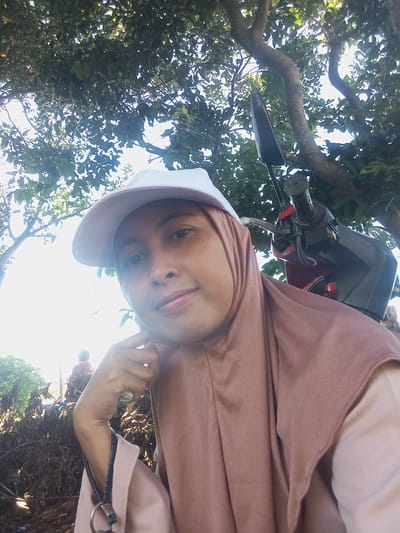 A woman wearing a hijab and baseball cap is taking a selfie. She is sitting on a red and black motorbike. The background includes trees and foliage. The photo was taken in Waung, Indonesia.