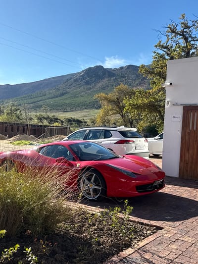 The image shows a red Ferrari parked on a brick driveway, next to a building labeled "OFFICE." Behind the Ferrari is a white Toyota. In the background, a mountain range is visible under a blue sky. The location is in Stellenbosch Nu, South Africa. The scene appears to be daytime and outdoors. No people are visible.