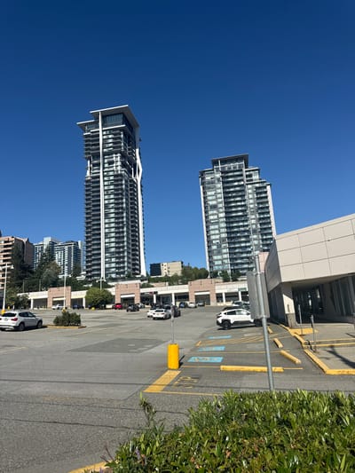 The image captures a bright, sunny day in Burnaby, Canada, with a clear blue sky. In the foreground, a paved parking lot with yellow painted lines and wheel stops is visible. Several cars are parked and driving in the lot, including a white SUV on the left, a white hatchback in the middle, and a white sedan on the right.

Dominating the midground are two modern, high-rise residential buildings. The building on the left is taller and features a distinctive white, angular facade element running up its side. The building on the right is slightly shorter and has a more conventional, stepped architectural design. Between and in front of these towers is a low-rise commercial building with a light-colored facade and storefronts. Signs visible on the commercial building include "Shore" and "Massage Therapy Cleaners".

In the background, other apartment buildings and trees can be seen, indicating a densely populated urban area. The overall scene depicts a typical urban environment with residential and commercial infrastructure.