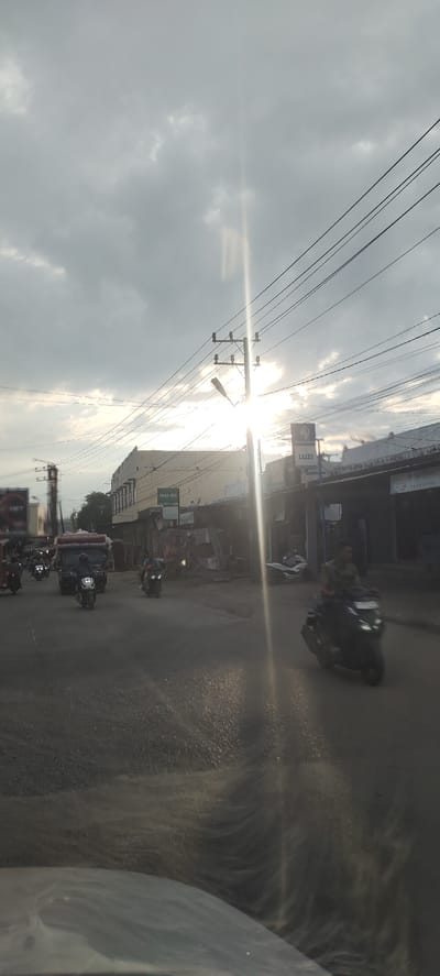 The image depicts an urban street scene in Keudegeudong, Indonesia, during what appears to be late afternoon or early evening, characterized by strong backlighting. The sky is mostly covered with scattered grey clouds, and intense sunlight projects a prominent lens flare and visible rays from the upper right side of the frame.
Multiple black power lines crisscross the upper and middle portions of the image, connecting to several utility poles. A street light fixture is mounted on the primary pole positioned towards the right of the center. On the left side of the street, a multi-story building, light in color, features a sign with "AWAN CELL" prominently displayed. Further down the street on the right, another commercial building has a sign that reads "LUZI."
The asphalt road surface in the foreground and midground exhibits a reflective quality, suggesting dampness or the effect of the low sun angle. Vehicular traffic includes numerous motorcycles and one small truck. A truck loaded with bulky items, possibly mattresses or textiles, is visible in the middle-left, preceding a person on a motorcycle. On the right side of the road, a person wearing a camouflage-patterned shirt is riding a dark-colored scooter, moving away from the viewer. Additional motorcycles and individuals are observed farther down the street. The perspective indicates the photograph was taken from inside a vehicle, with reflections visible along the bottom edge of the image.
