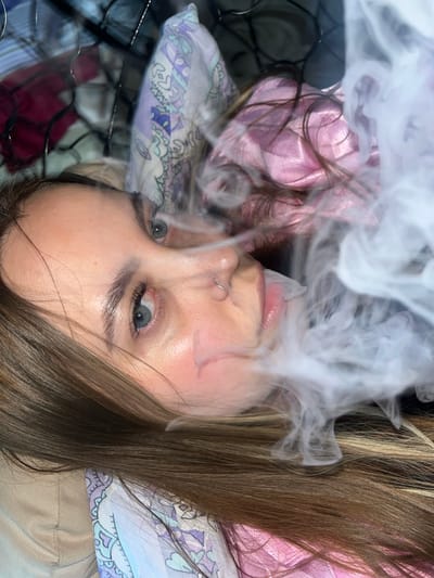 A young woman with brown hair and blue eyes is lying down, looking up at the camera. She has a nose ring in her left nostril. Smoke obscures part of her face. She is wearing a pink top and is lying on patterned pillows on a black wire rack. There is no evidence to support a location in Novopokrovskaya, Russia.