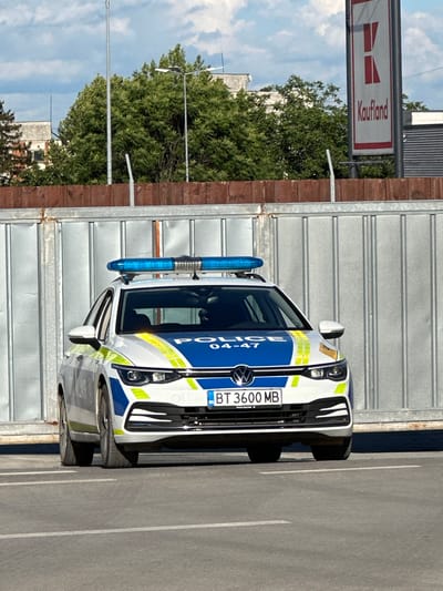 The image shows a white Volkswagen Golf police car with blue and yellow markings parked in an outdoor paved area. The car has a blue light bar on its roof and a license plate that reads "BT 3600 MB". The car is facing forward, and its headlights are on. In the background, there is a corrugated metal fence, trees, a sign for "Kaufland", and a cloudy sky. The scene appears to be in a parking lot, possibly near a commercial area. The location is identified as Gorna Oryahovitsa, Bulgaria.