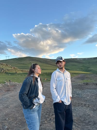The image shows a man and a woman standing outdoors in Hrazdan, Armenia. The background consists of a grassy mountain range meeting a gravel-covered area. The sky is visible, displaying a blue sky with fluffy white clouds. The man is wearing a light-colored pinstripe shirt, dark jeans, and a baseball cap. The woman is wearing a denim jacket over a light-colored hoodie and blue jeans.