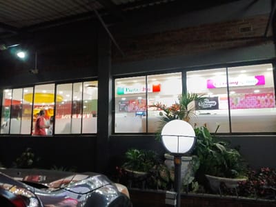 This nighttime image captures the brightly lit facade of a commercial building, likely located in Marondera, Zimbabwe. The scene features multiple storefronts observed through large glass windows, contrasting with the dark exterior of the building.

On the left, an establishment with a vibrant interior, featuring yellow and red decor, is visible. Several individuals, appearing to be customers or staff, are inside, engaged in typical store activities. Faintly visible on an interior wall are the words "LOVE LOVE."

To the right, two prominent business signs are displayed: "Pizza Inn," characterized by its green logo, and "Creamy Inn," identified by its pink logo, which appears twice. These are recognizable fast-food and ice cream chain establishments common in the region.

In the foreground, a spherical white outdoor lamp post casts light on an arrangement of potted plants, showcasing large green leaves and smaller reddish foliage, suggesting a landscaped area. A portion of a dark-colored car is visible in the bottom-left corner, indicating a nearby parking space. The time of day is clearly night, underscored by the dark sky and the illuminated interiors and outdoor lighting. The overall impression is that of a bustling commercial center operating after dark.