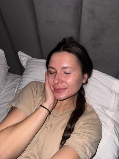 A young woman is asleep on a bed. She is lying on her side with her head resting on her hand. She has dark hair that is braided. She is wearing a tan T-shirt and a black bracelet. The bed has white sheets and pillows. The background is a gray upholstered headboard. The location is Akulovo (Gp Kubinka), Russia.