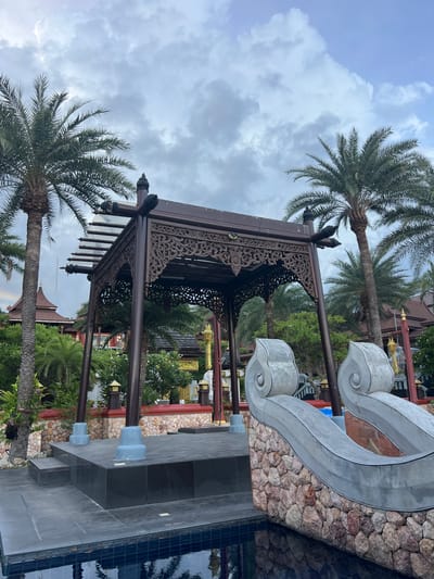 In Baan Lamai, Thailand, a wooden pavilion with intricate carvings stands on a raised platform adjacent to a swimming pool. The pavilion's roof is partially visible, supported by dark wooden posts. In the foreground, a stone wall forms the edge of the pool, topped with decorative gray elements resembling large shells or scrolls. Beyond the pavilion, palm trees and lush greenery are abundant, with traditional Thai-style buildings featuring distinctive pointed roofs visible in the background. The sky is cloudy, suggesting an overcast day.