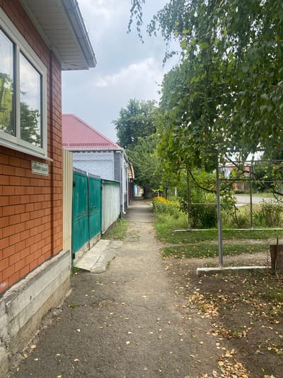The image depicts a narrow paved pathway running between two residential properties in the city of Maykop, Russia. The pathway is bordered on the left by a red brick house with a white-framed window and a concrete foundation. Adjacent to the brick house is a tall, green metal gate and a lighter colored fence. On the right, the pathway is bordered by a garden area with a metal structure, dense foliage, and yellow flowers. Fallen leaves are scattered across the pathway and garden. In the background, more residential buildings and trees are visible under an overcast sky. A street sign with Cyrillic text is affixed to the brick house. The scene suggests a quiet, suburban street.