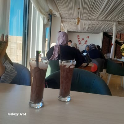 Two tall glasses of chocolate milkshake sit on a light-colored wooden table in the foreground. The glasses are filled with a dark brown liquid and have condensation on the outside. Black straws extend from each glass. In the background, several people are seated at tables, mostly women wearing headscarves. The setting appears to be an indoor cafe or restaurant with large windows that let in natural light. The ceiling is covered with white fabric, and a light fixture hangs from it. The overall ambiance suggests a casual gathering.