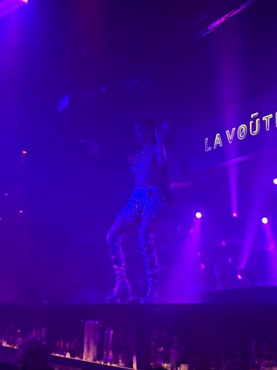 The image depicts a nightclub scene in Montreal, Canada, bathed in vibrant purple and pink lighting. A performer, dressed in a sparkly blue outfit and knee-high boots, is the central focus, captured mid-dance. The background features neon signage that reads "LA VOUTE," a prominent club in Montreal. Several spotlights cast beams of light through the hazy atmosphere, illuminating the stage and the performer. Below the stage, a bar area is visible, with bottles and glasses suggesting a lively atmosphere. The overall impression is of a dynamic and energetic nightlife setting.