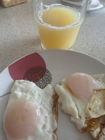 The image shows a close-up view of a breakfast setting. Two fried eggs with visible yolks are served on a white plate with a circular, abstract design. Next to the plate, a glass of yellow liquid, likely orange juice, is tilted, with bubbles on the surface. The eggs appear to be sunny-side up, with soft, pinkish yolks and cooked whites. The plate has a simple, modern design. The background consists of a speckled countertop. The lighting suggests it is daytime. The setting is a kitchen or dining area, typical of a home environment. There is no visible text in the image.