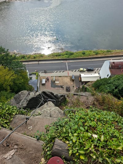 This image captures a high-angle view of a steep, rocky incline overlooking the Elbe River in Děčín, Czechia. The foreground shows a weathered wooden structure and dense green foliage. A steel cable and metal handrails are bolted into the rock face, indicating a hiking or climbing path descending towards a paved area below.

On the paved area, a group of people are gathered near a white truck with "BERNARD" printed on its side. Two yellow and black cars are parked on an adjacent road. Further beyond the road, the Elbe River is visible, reflecting the sunlight. A small, green-ringed object, possibly a buoys or a person in a life jacket, is in the river. A lone cyclist is seen on the road in the distance. The environment appears to be a scenic overlook or a recreational area.