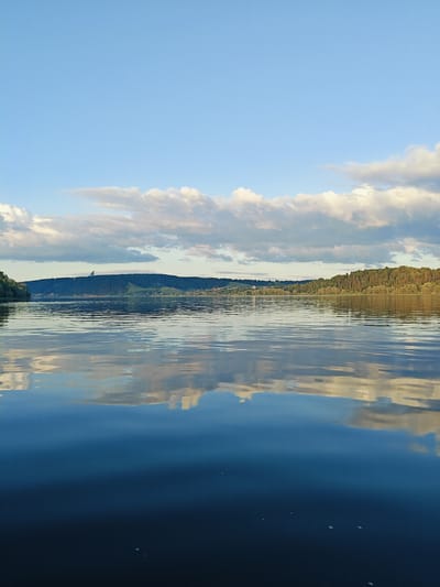 The image depicts a wide, calm river or lake in Ol'khovskoye, Russia. The sky is a clear blue with scattered, fluffy white clouds, some of which are illuminated by sunlight. The clouds are reflected in the still water, creating a mirror image. On the horizon, there is a lush, green forested hill with some scattered houses visible. The water surface is smooth with gentle ripples, reflecting the sky and the distant landscape. The scene conveys a sense of tranquility and natural beauty.