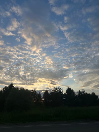 The image depicts a silhouetted line of trees and vegetation against a sky filled with scattered clouds. The clouds are illuminated by sunlight, creating a mix of light and dark patches. The bottom of the image shows a dark, indistinct strip that appears to be a road or pathway. There are no people or discernible structures visible. The scene is likely captured at dusk or dawn, given the lighting conditions. The location is identified as Povarёnki, Russia.