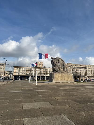 The image depicts the Place de l'Hôtel de Ville in Le Havre, France, under a partially cloudy blue sky with scattered white cumulus clouds. The foreground consists of extensive, light grey rectangular paving stones.
The central subject is the Le Havre War Memorial, a large, monumental stone sculpture situated slightly to the right of the frame's center. It features multiple human figures sculpted in relief on a substantial base, which bears the inscription "1939-1945." To the left of the memorial, three white flagpoles are present. The rightmost flagpole (closest to the memorial) displays a French tricolor flag (blue, white, red vertical stripes) at full mast, while the middle flagpole flies a French tricolor flag at half-mast. The leftmost flagpole is bare.
A single individual, dressed in dark clothing, is observed walking from left to right across the mid-ground of the plaza. In the background, distinctive multi-story modernist buildings, characteristic of Le Havre's post-war reconstruction, line the square. These structures feature light-toned facades with numerous repetitive windows and ground-floor commercial establishments. Several black, multi-armed streetlights are visible on the left side of the scene. Sparse vehicular traffic is present on distant roads bordering the plaza. The scene is illuminated by natural daylight.