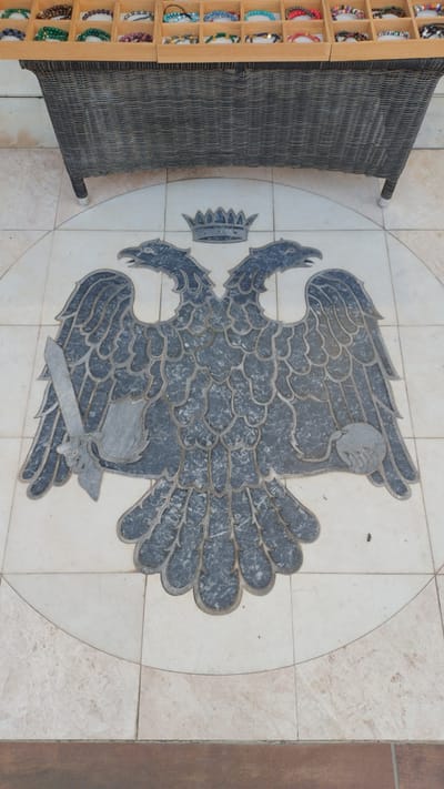 This is a photograph of a decorative stone inlay of an eagle with spread wings, a crown, and a sword. The eagle is depicted in dark gray stone against a lighter, cream-colored tiled background. The inlay is circular, with the eagle positioned in the center. To the left of the inlay is a dark, woven bench or chair. To the far left of the image is a display stand filled with colorful bracelets. The setting appears to be outdoors, possibly a patio or promenade, given the tiling and the presence of outdoor furniture and a retail display. The lighting suggests it is daytime, with no strong shadows indicating a particular time of day, and the weather appears to be clear. The image is a still shot, with no discernable activity taking place. There is no visible text in the image. The overall impression is one of a decorative feature in a public or commercial space.