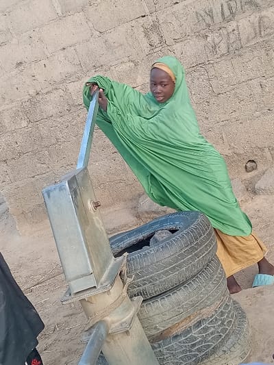 In Ayama, Nigeria, a young female operates a manual well pump. She wears a light green hijab over a mustard yellow garment, with one foot bare and the other in a light blue slipper. Her hands firmly grasp the silver-grey metal pump handle. The pump's base is stabilized by straps attached to a stack of three worn black car tires, with a visible rock inside the topmost tire. The immediate ground consists of dry, light brown earth. A textured wall of light brown concrete blocks or bricks forms the background, showing signs of age and minor surface markings.
