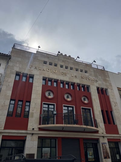 The image depicts the "Palacio de la Música" (Palace of Music) building, located in Buñol, Spain. The structure features a multi-story facade constructed with light-colored, possibly beige or off-white, stone cladding and prominent reddish-brown sections.
The upper portion of the building displays the golden lettering "PALACIO DE LA MUSICA" horizontally across the stone facade, beneath a row of small, rectangular windows. Above this, a flat roofline is visible, topped with a dark metal railing and several black outdoor light fixtures.
The middle section of the facade is primarily reddish-brown with vertical architectural elements and dark-framed windows. Three circular recesses on this level each contain a dark, weathered bust, facing forward. Below these busts, a curved balcony with a black metal railing extends outward, situated in front of a pair of dark-framed double doors. Additional dark-framed windows are present on either side of this central section.
The ground level features dark roll-down shutters covering storefronts, along with glass doors and display windows containing visible posters. A dark metallic structure, possibly a railing or part of an entrance ramp, is partially visible at the bottom left of the frame.
The sky overhead is largely overcast with significant cloud cover, but a bright sunspot is visible behind the clouds, creating a slight glare. A thin, dark wire with a small, indistinguishable object attached to it spans diagonally across the upper left portion of the sky. No individuals are present in the scene.