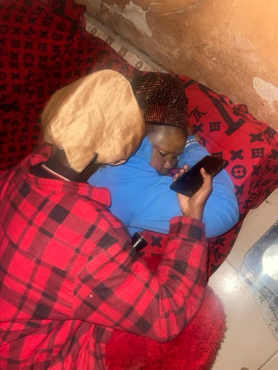 Two individuals are pictured reclining on a bed draped with a red blanket adorned with black Louis Vuitton-style symbols. One person, clad in a blue shirt and a jeweled head covering, holds a phone. Another person, wearing a red and black plaid jacket and a tan hat, is positioned in front. A fluffy red rug is on the right, while the wall shows signs of wear and discoloration. A shiny metallic object is also visible at the top of the image.