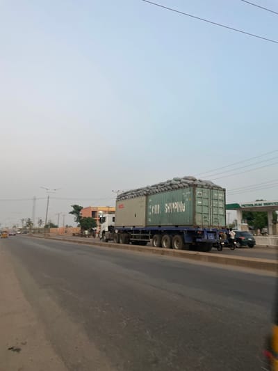 A slightly tilted, medium-shot captures a commercial truck stopped on a paved road in what appears to be Hayin Banki, Nigeria, based on visual characteristics of the built environment. The truck is carrying two stacked, green cargo containers filled with what appear to be bagged goods, stacked high over the top of the shipping containers. The bags on top of the containers appear to be covered with light-colored fabric. A motorcycle is beside the truck with two riders. Buildings, vegetation, and power lines are visible along the roadside. The sky is overcast and the environment shows potential signs of atmospheric haze or dust. The road surface is worn and shows signs of use. There are a number of buildings and signs of commercial activity nearby.