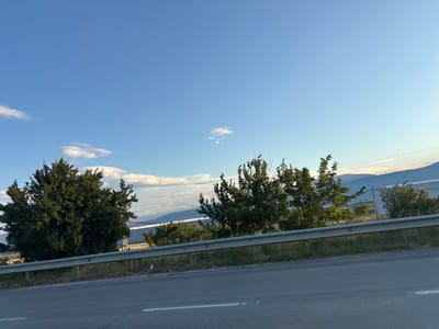 This media file depicts a roadside scene in Dzveli Kanda, Georgia, under a clear blue sky with scattered white clouds. The foreground is dominated by a grey asphalt road with a white lane marking. A metal guardrail runs along the side of the road, separating it from a grassy verge with some litter. Beyond the verge, there are trees with lush green foliage. In the middle ground, several buildings with white facades are visible, and in the distance, rolling hills and mountains can be seen under the soft light of either early morning or late afternoon. The overall impression is one of a peaceful, rural landscape. There are no people or specific activities visible, but the scene suggests a moment of travel or observation from a moving vehicle. The weather appears to be pleasant, with good visibility.