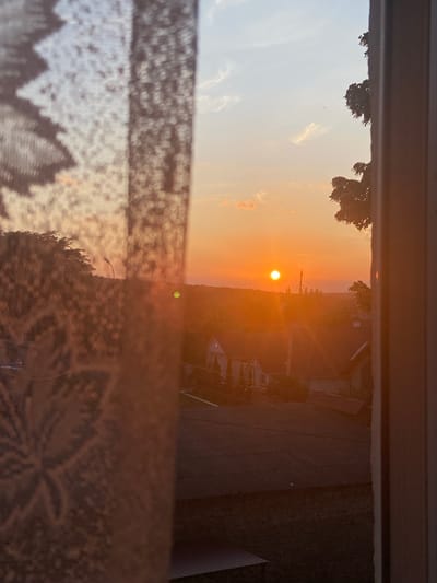 The image captures a sunset viewed through a window with a sheer lace curtain in the foreground. The sun, a bright orange orb, is low on the horizon, casting a warm glow over a semi-urban landscape. The scene appears to be from Soroca, Moldova, given the architectural style of the houses and the general appearance of the surroundings. The curtain, partially obscuring the view, has a floral pattern. In the mid-ground, rooftops of houses are visible, some with tiled roofs. Beyond the houses, trees and a streetlamp can be seen. The sky is a gradient of orange and pale blue, with a few wispy clouds. A tall structure, possibly a utility pole or communication tower, is visible in the distance. The overall impression is one of a peaceful, late afternoon or early evening in a residential area.