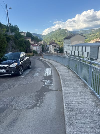 The image depicts a curved road with a concrete barrier and metal railing on the right. White markings are visible on the road surface. Two cars are parked on the road, one is a dark SUV, and the other is a black car. In the background, a town or village is nestled on a hillside, with buildings of various architectural styles and colors. Lush green trees cover the slopes, and in the distance, a body of water can be seen, with mountains rising beyond it. The sky is a clear blue with some fluffy white clouds. A lamppost and electrical wires are visible on the left side of the image, suggesting an urban or semi-urban environment. The overall scene suggests a sunny day in a scenic location. The license plate of the dark SUV is partially visible and reads "881-XY".