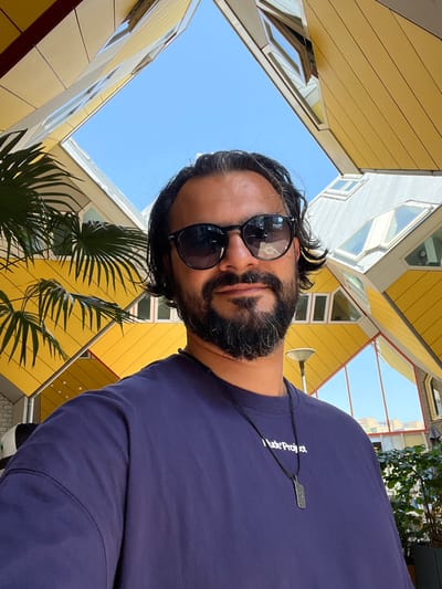 A man with dark hair and a beard is taking a selfie in front of the Cube Houses in Rotterdam, Netherlands. He is wearing sunglasses and a dark blue t-shirt with a white inscription and a pendant necklace. The Cube Houses, with their distinctive yellow and white angular facades, are visible in the background, along with a clear blue sky. A palm tree is partially visible on the left side of the image.