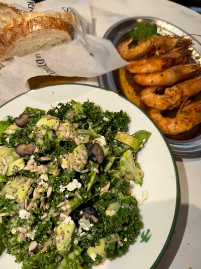 The image shows a table setting with three food items. On the left is a dish of large, grilled shrimp with a dark reddish seasoning and an oily base. To the right, a white bowl with a green rim contains a salad of kale, avocado, blueberries, sunflower seeds, and feta cheese, dressed with a light-colored dressing. At the lower left is a basket lined with tissue paper holding slices of bread. The environment suggests a restaurant setting. The text on the tissue paper indicates "LE DITRI," potentially a brand or location. This image was likely taken in Tirana, Albania.