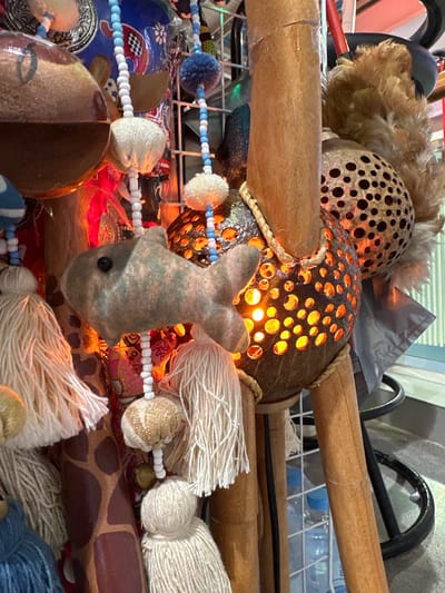 This media file showcases a close-up view of a collection of decorative objects, likely part of an art installation or a display in a shop. The main subjects are intricately carved coconut shells, serving as lamps or lanterns, with the warm glow of internal lights shining through numerous circular perforations. These illuminated shells are interspersed with various textural elements, including a small, fabric fish with a black bead eye, bundles of natural fiber tassels in shades of cream and white, and sections of bamboo. Decorative beads, in blue, white, and clear, string together some of these elements, creating a sense of handcrafted artistry. The background is a blur of colors and textures, hinting at a busy environment with other decorative items and possibly some metal shelving. The lighting suggests an indoor setting, possibly illuminated by the internal lights of the coconut shells, creating an ambiance that is warm and inviting. The overall impression is one of artisanal creativity and a blend of natural materials with decorative embellishments. There are no visible people or specific location cues beyond the general context of Wichit, Thailand. The time of day and weather are not discernible from the image. There is no visible text.