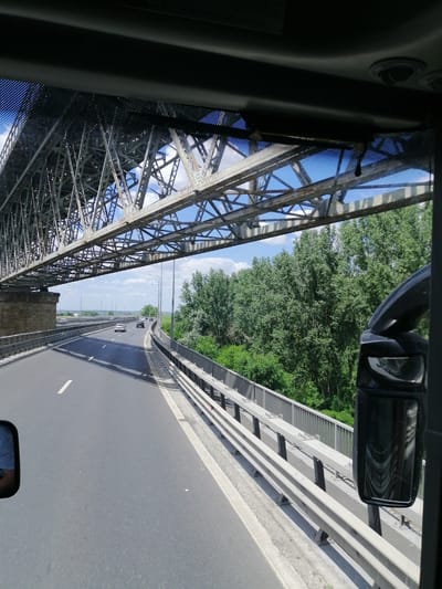 A wide-angle, exterior shot captures a view from a vehicle driving on a highway beneath a large, steel truss bridge. The bridge spans across the road and stretches into the distance, with its intricate metal structure dominating the upper portion of the frame. Sunlight streams through the open framework of the bridge, casting shadows on the asphalt road below.

The road is paved and appears to be in good condition, with a single white line marking the lane. Several cars are visible driving ahead on the highway, traveling in the same direction as the camera. The highway is bordered by a metal guardrail on the right side.

To the right of the road, dense green trees and foliage form a thick treeline, suggesting a natural or rural environment. The sky above is a clear blue with scattered white clouds.

In the foreground on the right side of the image, a large, black side-view mirror is prominently featured, reflecting the surrounding scenery, including the bridge and trees. On the left side of the frame, a portion of a person wearing a blue shirt is visible in the reflection of a smaller mirror, suggesting the photograph was taken from inside a vehicle.

The scene is set in Stelnica, Romania, under a bright, sunny sky.