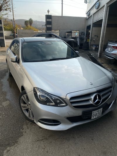 The image presents a silver Mercedes-Benz E-Class sedan (W212 facelift model) parked on a wet, dark asphalt surface, indicative of a recent wash, at a facility in Ma'raba, Syria. The vehicle's Syrian license plate, displaying "517 SYR 9920," is mounted on the front bumper. The car features a dark-colored roof, potentially a panoramic sunroof.

To the right of the Mercedes, a light-colored building with two garage bay doors, one partially open, identifies an auto service or car wash establishment. Two clocks are affixed to the building's facade above the bays. Inside the open bay, a stack of tires is visible. The rear section of a grey sedan is partially visible to the far right.

In the mid-ground, a concrete wall with a decorative pillar topped by an outdoor lantern light fixture is present. Beyond this wall, another building with small rectangular windows and a darker vehicle are discernible. The distant background shows hills with sparse vegetation, including palm trees, under a partly cloudy sky. Overhead power lines are visible in the upper left quadrant. No individuals or specific actions are depicted within the scene.