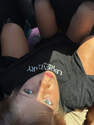 A young child lies with their head tilted up towards the camera, a black t-shirt with "LEGENDARY" printed on it across their chest. The child has fair skin, light hair, and striking blue eyes. Part of their face is shadowed by sunlight. One leg is extended upwards and visible next to the t-shirt. A stuffed animal, possibly purple, is visible in the lower left corner. The background is mostly dark, suggesting they are indoors, possibly in a car. The precise location in Kunje, Montenegro cannot be determined from the image.