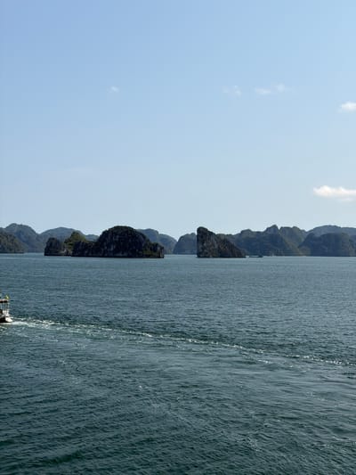 This landscape image depicts a serene and majestic bay, consistent with the characteristic scenery of Ha Long Bay in Ha Long Ward, Vietnam. The scene is divided horizontally into three main sections: the clear sky, the dramatic limestone islands, and the vast expanse of water.
The foreground and mid-ground are dominated by the dark blue-green waters of the bay. The surface of the water is textured with numerous ripples, reflecting sunlight in shimmering patterns. A distinct, foamy white wake trails diagonally across the water from the bottom right, indicating the movement of a boat. Only a small portion of the boat, likely its stern or side with a railing and what appears to be a yellow flag or banner, is visible in the bottom right corner of the frame.
Along the horizon, a series of imposing limestone karst islands rise sharply from the water. These islands are covered in dense, dark green foliage, contrasting with the lighter rock formations that are also visible. They vary in size and proximity, creating a sense of depth and an undulating, rugged silhouette against the sky.
The sky, filling the upper portion of the image, is a clear, light blue with a few faint, wispy white clouds scattered across it. The overall brightness and clarity suggest it is daytime, likely late morning or early afternoon, under fair weather conditions. There is no visible text in the image.