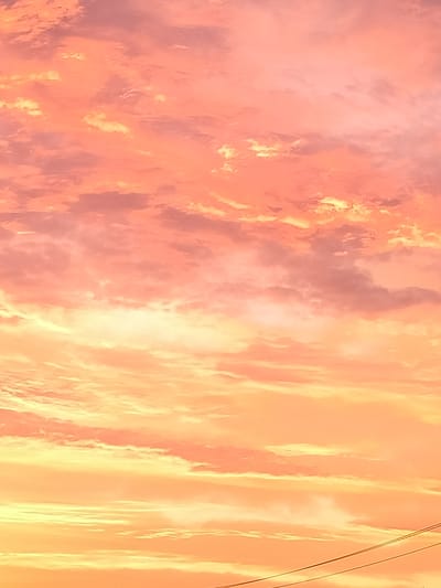 The image captures a sky filled with vibrant orange and yellow clouds, suggesting a sunset or sunrise. The clouds are varied in texture and density, with some appearing wispy and others more voluminous. The colors blend seamlessly, creating a warm and atmospheric scene. In the lower right corner of the image, two parallel wires, likely part of an electrical or telephone line, are visible against the bright sky. The rest of the image is dominated by the sky and clouds, with no discernible ground or structures. The overall impression is one of natural beauty and tranquility, with the man-made wires providing a subtle contrast to the expansive natural canvas. This image is of the sky above Yashkino, Russia.