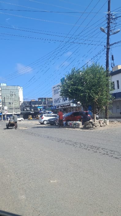 The image depicts an urban street scene in Majengo ward, Kenya, under a clear blue sky. Numerous utility wires crisscross overhead.

In the immediate foreground, an asphalt road surface is visible, showing tire marks and minor debris. Towards the left midground, a man in a dark shirt and trousers pushes a two-wheeled handcart, moving from left to right across the street. Behind him, several parked vehicles, including a white sedan and a dark sedan, line the far side of the road. Further left, a modern multi-story building with a reflective facade stands. Several auto-rickshaws ("tuk-tuks") are visible in the background street.

On the right midground, a prominent green leafy tree grows on a small unpaved median. Beneath the tree, two male individuals are present: one man in a red shirt stands, facing right, while another man in a dark shirt appears to be seated or squatting. Adjacent to these individuals, multiple paint cans of various colors are stacked on the ground, alongside loose construction materials such as stones or bricks. A dark red SUV is parked next to this grouping, with another dark vehicle partially visible behind it. A dark utility pole, heavily laden with wires and featuring two streetlights, rises vertically on the far right.

The background consists of several multi-story buildings, including a distinctive white building with dark blue trim on the right and other structures with varied architectural styles and visible commercial signage. The scene is illuminated by daylight, indicating active city life.