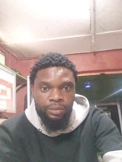 A close-up shot showcases a dark-skinned man with short, curly hair and a full beard. He is wearing a dark hoodie with a light gray hood lining. His expression is neutral, with direct eye contact. The background is a dimly lit room with a computer monitor visible on the left side. The lighting in the image creates a slightly grainy and muted color palette.