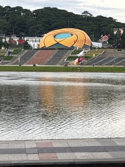 The image shows the Xuan Huong Lake in Da Lat, Vietnam, on an overcast day. In the foreground, the rippling surface of the lake reflects the sky and surrounding landscape. Beyond the lake, a grassy embankment leads up to a wide, terraced walkway. This walkway is lined with steps and planters, and several individuals are visible strolling along it. Dominating the mid-ground is a large, distinctive building with a golden, petal-like roof structure, featuring a prominent blue circular skylight. Behind this building, a dense forest covers a hillside, with several houses and buildings interspersed among the trees. The overall scene depicts a public recreational area with notable architectural features and natural scenery.