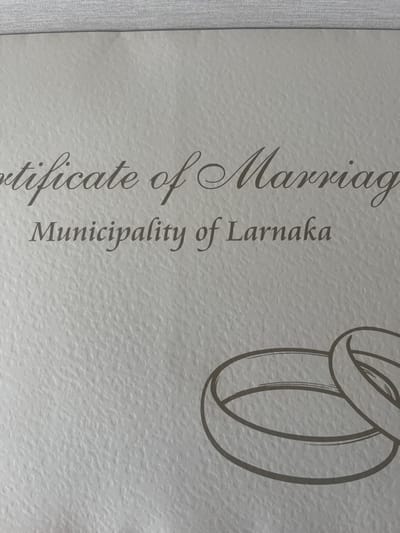 A close-up, slightly angled photograph depicts the upper portion of a light-colored, textured document, identified as a Certificate of Marriage. The document prominently features the partial text "tificate of Marriag" in an ornate serif font and "Municipality of Larnaka" in a plainer serif font, both printed in a dark grey-brown color. The bottom-right section of the visible document includes an illustration of two interlocked wedding rings, rendered in the same dark grey-brown. The paper exhibits a fine, irregular texture. The upper edge of the document is slightly blurred, resting on or against a lighter, possibly fabric surface. This scene is located in Agios Tychonos, Cyprus.