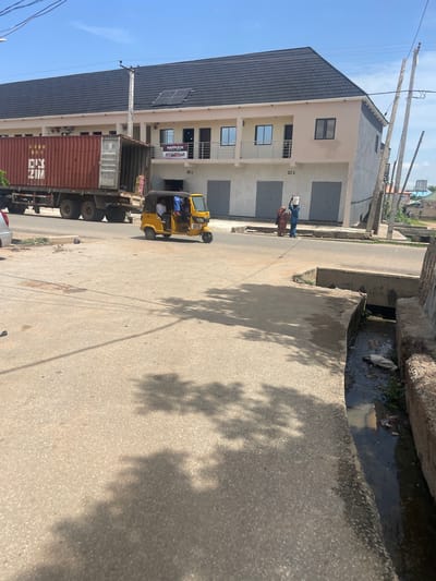 This media file captures a street scene in Jimeta, Nigeria, likely during the daytime under a clear sky. The image is oriented vertically, showing a street lined with buildings and a drainage channel.
In the foreground, a large truck carrying a shipping container is parked on the right side of the road. Next to it, a yellow auto-rickshaw, also known as a "Keke Napep," is stopped, with several passengers visible inside. The rickshaw has text on its side, but it is not clearly legible.
Further down the street, two individuals are walking. One is carrying a white bucket on their head, and the other is dressed in a patterned fabric. Behind them, a multi-story building with a dark-tiled roof and light-colored walls stands prominently. This building features several windows and balconies, and what appears to be a sign with the text "HAPPULCH" and an image of a woman. A solar panel is affixed to the roof of this building.
The street is paved with asphalt, and a concrete drainage channel runs alongside the road, containing murky water and some debris. The shadows of trees are cast across the road and the drainage channel, suggesting the presence of nearby foliage and the time of day. The overall scene depicts a typical urban street environment in Nigeria, with everyday activities of transportation and pedestrian movement.