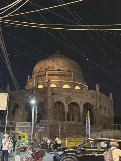 This is a nighttime photograph of a historical monument in Delhi, India, possibly a tomb, characterized by a large dome and arched openings. The monument is constructed of stone and appears aged, with visible wear and tear. Artificial lighting illuminates the interior of the arched spaces, casting a warm glow. Overhead, a complex network of electrical wires crisscrosses the dark sky, indicating an urban setting.

In the foreground, there is street activity. A yellow auto-rickshaw is parked, and several people are standing around it. One person, wearing a white t-shirt and a cap, is holding a phone. Another person, in a green t-shirt, is also looking at their phone. A red motorcycle is parked in front of them. Further to the right, a black car is visible, and two people are on a motorcycle. The overall environment suggests a lively urban street scene at night, with the historical monument serving as a prominent backdrop.
