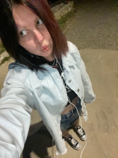 A selfie captured at dusk. The subject, a young person with auburn and black hair, is facing the camera with pursed lips. They are wearing a light-colored denim jacket over a dark top with visible lettering, a denim skirt, and black-and-white Converse sneakers. A white earphone wire is visible. The background is a concrete or asphalt surface. The location is not identifiable as Kunje, Montenegro, from visual information.