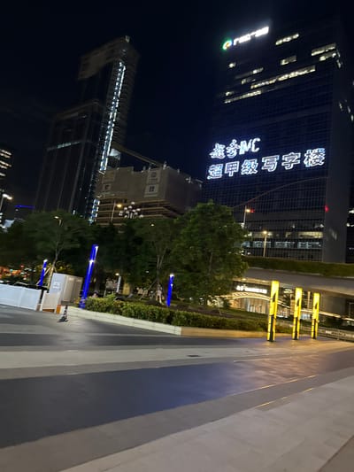 This image depicts a nighttime view of a modern urban landscape in Huangpu District, China. In the background, two prominent skyscrapers dominate the skyline. The taller building on the left appears to be under construction, with visible scaffolding and unfinished sections. The building on the right is a completed high-rise with illuminated windows and a large, bright sign displaying Chinese characters and "MC" in English. The sign reads "嘉信MC 超甲级写字楼," which translates to "Jiaxin MC Super Grade Office Building."
In the foreground, a paved walkway with distinct patterns of dark gray, light gray, and blue sections leads the eye towards the buildings. To the left of the walkway, there are white barricades and portable restroom units. Several tall, slender, blue-lit poles are strategically placed along the edge of the walkway, providing ambient lighting. Lush green trees and manicured bushes are visible between the walkway and the buildings, adding a touch of nature to the urban environment. Further into the mid-ground, a series of tall, yellow-lit cylindrical structures stand out, illuminating the area beneath an elevated walkway or bridge. The overall scene conveys a sense of modernity and urban development.